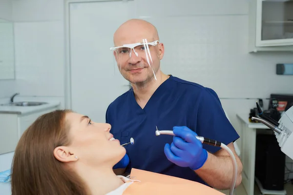 A kind dentist in a medical face shield holds a dental mirror and drill near a lying young woman in the dental chair. A doctor and his female patient during treatment in a dentist\'s office.