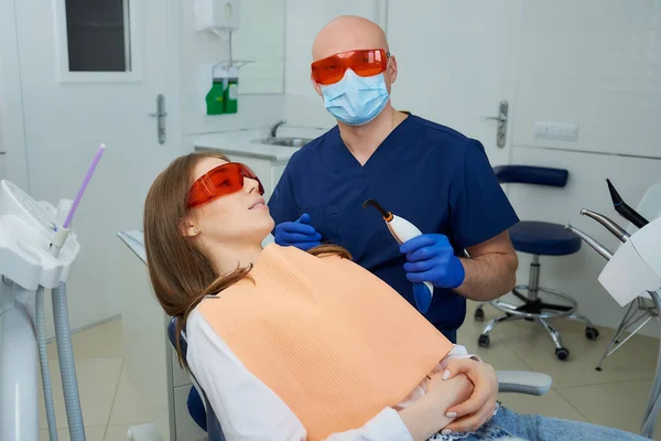 A dentist in a medical face mask and UV protection glasses holding a dental polymerization light near a girl in the dental chair. A doctor and a female patient during treatment in a dentist\'s office.