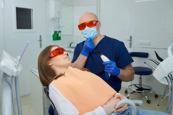 Dentist in UV protection glasses is checking a medical face mask and holding a dental polymerization light near girl in the dental chair. Doctor and female patient during treatment in dentist\'s office