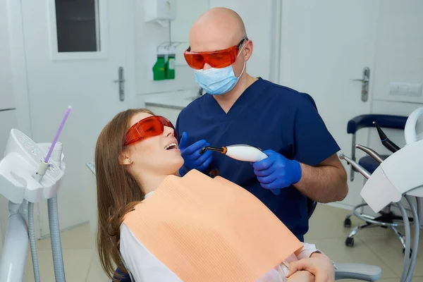 A dentist in a medical face mask and UV protection glasses holding a dental polymerization light near a woman in the dental chair. A doctor and a patient before treatment in a dentist\'s office.