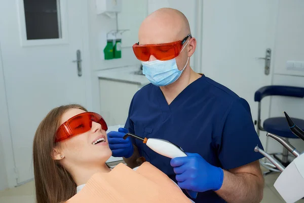 A dentist in a medical face mask and UV protection glasses holding a dental polymerization light near a young woman in the dental chair. A doctor and a patient started treatment in a dentist's office.