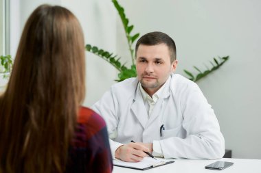 Beyaz laboratuvar önlüklü beyaz bir doktor bir masada oturuyor ve bir doktorun ofisinde kadın bir hastanın şikayetlerini yazıyor. Hastanede doktor randevusunda uzun saçlı bir kadın..
