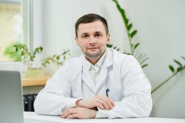 Beyaz laboratuvar önlüklü beyaz bir doktor hastanede kollarını kavuşturmuş oturuyor. Laptopun yanında bir terapist doktor muayenehanesinde hasta bekliyor..