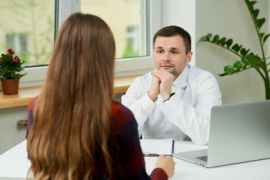Beyaz laboratuvar önlüklü beyaz bir doktor bir masada oturuyor ve bir doktorun muayenehanesinde bir kadın hastayı tedavi etmeye çalışıyor. Hastanede doktor randevusunda uzun saçlı bir kadın..
