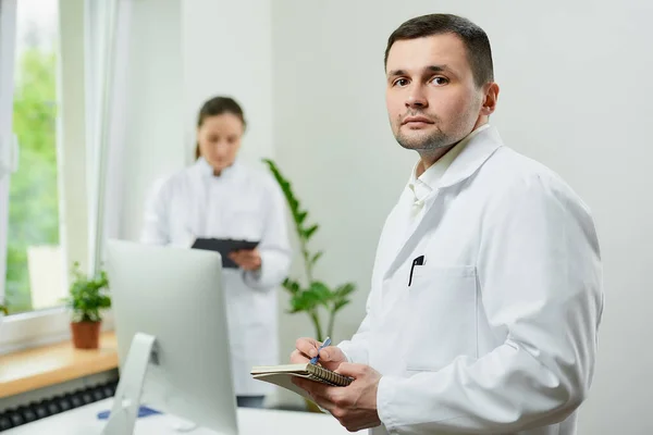 A serious doctor with a notebook and a pen is thinking about the patient's medical history in a hospital. A female therapist is doing notes in a clipboard near an all in one computer in a clinic.