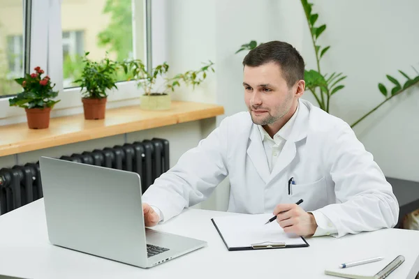A happy caucasian doctor in a white lab coat is listening to a patient on an online meeting on a computer in a hospital. A therapist is sitting at a desk with a laptop in a doctor's office.