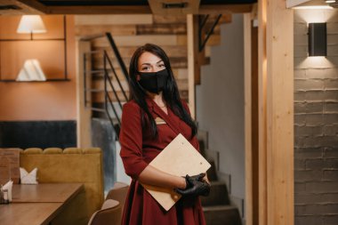 Siyah tek kullanımlık tıbbi eldiven giyen bir bayan restoran müdürü, elinde bir restoranda ahşap menü tutarken tıbbi maske takıyor. Yakut elbiseli, uzun saçlı bir kafe sahibi..