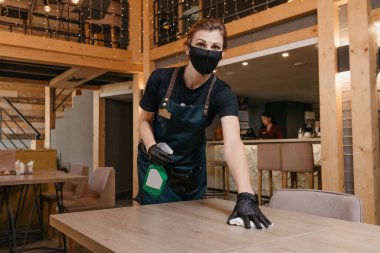 Siyah tıbbi maske takan ve tek kullanımlık tıbbi eldiven takan bir garson elinde dezenfektan ve restoranda bez parçasıyla masaları temizleyen bir şişe tutuyor. Kahveci bir kafede müşterilerini bekliyor..