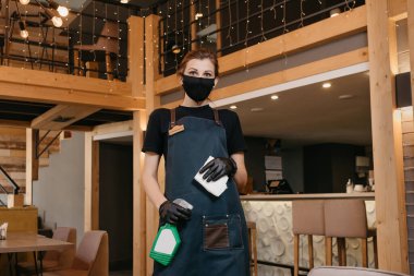 Tıbbi maske takan ve siyah tek kullanımlık tıbbi eldiven giyen bir garson restoranda masaları temizlemek için bir şişe dezenfektan ve bez tutuyor. Kahveci bir kafede müşterilerini bekliyor..