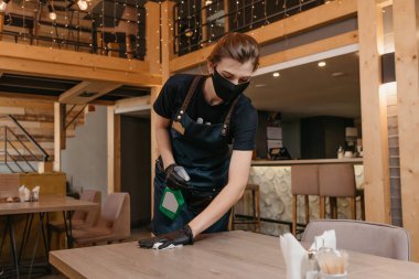 Siyah tıbbi maske takan ve tek kullanımlık tıbbi eldiven giyen bir garson elinde dezenfektanla bir şişe tutuyor ve restoranda bir bez parçasıyla masayı temizliyor. Bir barista müşteri bekliyor..