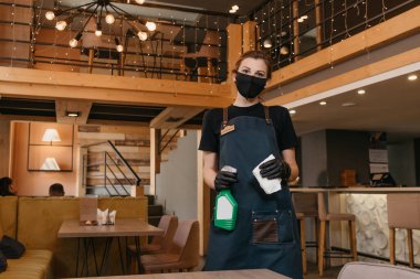 Siyah tıbbi maske ve tek kullanımlık tıbbi eldiven takan bir garson restoranda masaları temizlemek için bir şişe dezenfektan ve bez tutuyor. Kahveci bir kafede müşterilerini bekliyor..