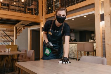 Siyah tıbbi maske takan ve tek kullanımlık tıbbi eldiven takan kibar bir garson restoranda içinde dezenfektan olan bir şişe ve bir bez parçasıyla masaları temizliyor. Personel müşteri bekliyor..