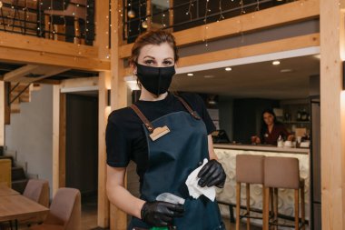 Önlük takan şık bir garsonun yakın portresi, siyah bir tıbbi maske ve tek kullanımlık eldiven, restoranda içinde dezenfektan ve beyaz bez bulunan bir şişe tutuyor..