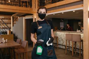 Önlük takan şık bir garson, siyah tıbbi maske takan ve tek kullanımlık tıbbi eldiven bir restoranda içinde dezenfektan ve bez bulunan bir şişe tutuyor. Personel bir kafede müşterilerini bekliyor.