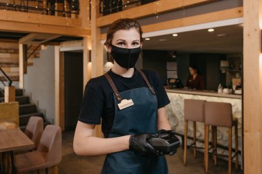 Önlük takan, siyah tıbbi maske takan ve tek kullanımlık eldiven takan bir garson restoranda kablosuz ödeme terminali tutuyor. Personel, müşterinin faturayı ödemesini bekliyor..
