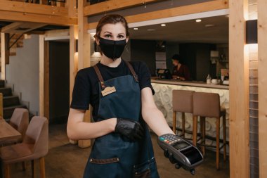 Önlük takan, tıbbi maske takan ve tek kullanımlık tıbbi eldiven takan bir garson restorandaki bir müşteriye kablosuz ödeme terminali veriyor. Personel müşterinin faturayı ödemesini bekliyor..