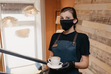 Siyah yüz maskesi ve tek kullanımlık eldiven takan bir garson restoranda kahve servisi yapıyor. Şirin bir barista bir kafede müşterilerini bekliyor..