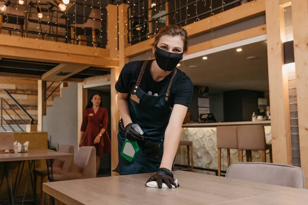 A kind waitress who wears a black medical face mask and disposable medical gloves is holding a bottle with sanitizer and cleaning tables with a rag in a restaurant. A barista is waiting for clients.