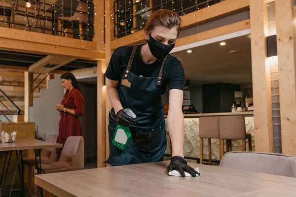 Siyah tıbbi maske takan ve tek kullanımlık eldiven takan sevimli bir garson bir restoranda dezenfektan ve bez parçasıyla temizlik masaları tutan bir şişe tutuyor. Personel bir kafede müşterileri bekliyor..