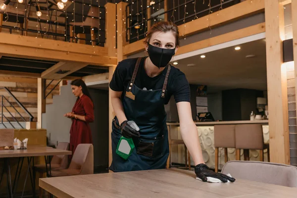 A waitress who wears a black medical face mask and disposable medical gloves is holding a bottle with sanitizer and cleaning tables with a rag in a restaurant. Staff is waiting for clients in a cafe.