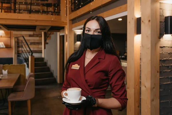 A gorgeous female restaurant manager who wears a black face mask and disposable gloves is holding a cup of coffee in a restaurant. A pretty cafe owner in a ruby dress is waiting for clients.