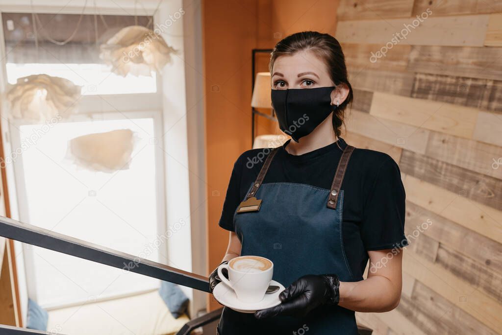A waitress who wears a black face mask and disposable gloves is serving a cup of coffee in a restaurant. A cute barista is waiting for clients in a cafe.