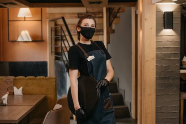 Tıbbi maske takan ve siyah tek kullanımlık tıbbi eldiven giyen bir garson bir restoranda plastik tepsiyle poz veriyor. Şirin bir barista bir kafede müşterilerini bekliyor..