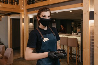 Önlük takan, siyah tıbbi maske takan ve tek kullanımlık eldiven takan sevimli bir garson restoranda kablosuz ödeme terminali tutuyor. Personel, müşterinin faturayı ödemesini bekliyor..