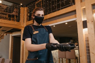 Önlük takan, tıbbi maske takan ve tek kullanımlık tıbbi eldiven takan bir garson restorandaki bir müşteriye kablosuz ödeme terminali veriyor. Personel, tüketicinin faturasını ödemesini bekliyor..