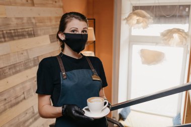 Siyah yüz maskesi ve tek kullanımlık eldiven takan bir garson restoranda kahve servisi yapıyor. Şirin bir barista bir kafede müşterilerini bekliyor..