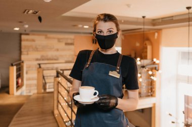 Siyah maske takan ve tek kullanımlık eldiven takan bir garson bir restoranın ikinci katında kahve servisi yapıyor. Şirin bir barista bir kafede müşterilerini bekliyor..