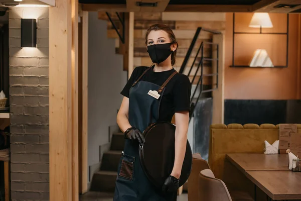 A waitress who wears a medical face mask and black disposable medical gloves is posing with a plastic tray in a restaurant. A cute barista is waiting for clients in a cafe.