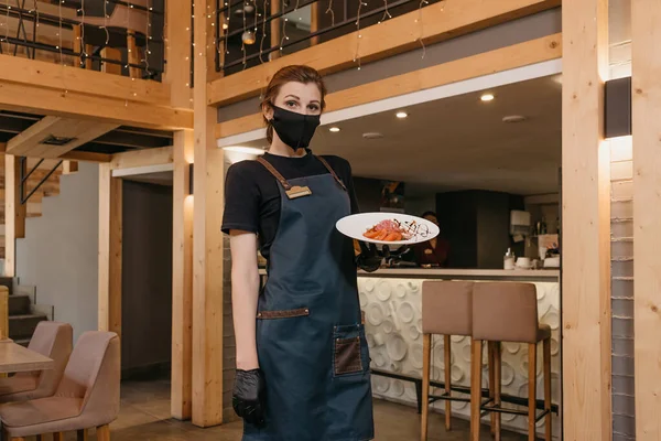 A pretty waitress who wears a black face mask and disposable gloves is holding a mint and dolendwitz salad in a restaurant. A cute barista is serving clients in a cafe.