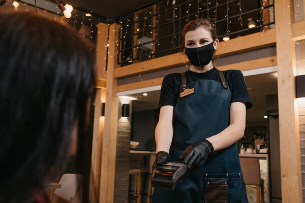 A waitress who wears a medical face mask and disposable medical gloves is handing a wireless payment terminal to a female client in a restaurant. The staff is waiting for the consumer to pay a bill.