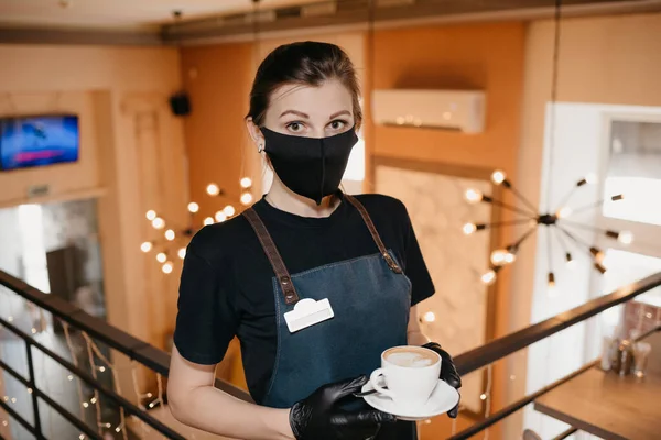 Siyah maske takan ve tek kullanımlık eldiven takan güzel bir garson bir restoranın ikinci katında kahve servisi yapıyor. Şirin bir barista bir kafede müşterilerini bekliyor..