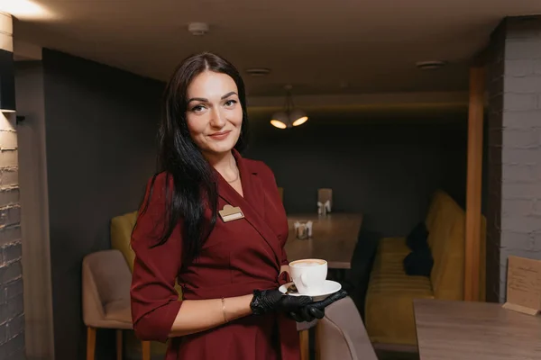 A female restaurant manager in black disposable gloves is serving a cup of coffee in a restaurant. A kind cafe owner in a ruby dress is waiting for clients.