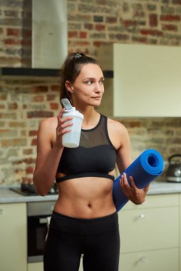 Siyah takım elbiseli gülümseyen sporcu bir kız, evde antrenmandan önce susuzluğunu gidermek için mavi yoga minderi ve beyaz plastik karıştırıcıyı tutuyor..