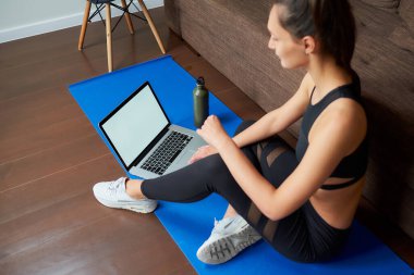 Siyah dar pantolonlu ve üstü mavi yoga minderinde elinde bilgisayar ve şişeyle oturan sportif bir kızın yan çekilmiş bir fotoğrafı. Bir dizüstü bilgisayar ve fitness kadınının maketi..