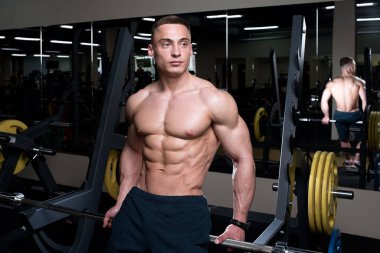 Üstsüz bir vücut geliştirici spor salonundaki halterin üzerinde oturuyor. Kafkas erkek fiziği sporcusu kaslı gövdesini gösteriyor..