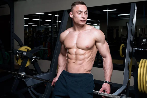 Soluk tenli üstsüz bir vücut geliştirici spor salonundaki bir halterin üzerinde oturuyor. Kafkas erkek fiziği sporcusu kaslı gövdesini gösteriyor..