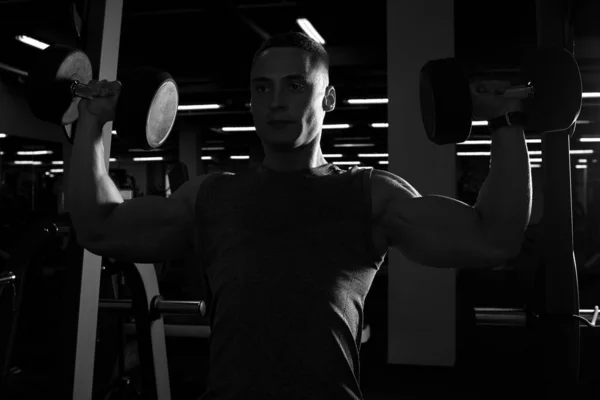 A muscular man is doing dumbbells overhead press in a gym. A bodybuilder is doing a workout with dumbbells for getting ready for the bodybuilding competition. Black and white silhouette photo.