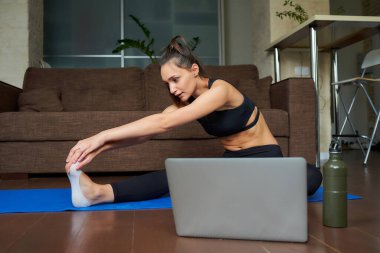 Siyah takım elbiseli sportif bir kız dizüstü bilgisayarında bir video ile esneme egzersizi yapıyor. Bir jimnastik koçu evdeki kanepenin yanındaki mavi yoga minderinde sanal fitness dersi veriyor.