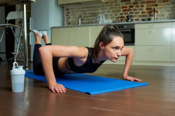 A sporty skinny girl in a black workout tight suit is doing knee push-ups to warming up pectoral muscles, triceps, and shoulders on the blue yoga mat at home.