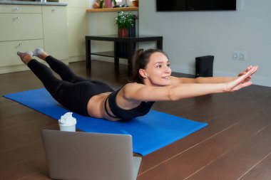 Siyah takım elbiseli gülümseyen sporcu bir kız klasik tekne egzersizini sırtı için yapıyor. Süpermen pozu veren bir koç evdeki mavi yoga minderinde uzaktan kumandalı bir beden eğitimi dersi veriyor..