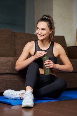 Siyah spor takım elbiseli sportif bir kız gülümsüyor ve evdeki koltuğun yanındaki mavi yoga minderindeki antrenman setleri arasındaki susuzluğunu gidermek için yeşil alüminyum bir şişe açıyor..