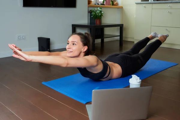 A smiling sporty girl in a black workout tight suit is doing the classical boat exercise for the back. A coach in a superman pose conducting a remote online fitness class on the blue yoga mat at home.