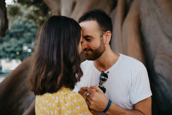 A  girl in a hat and a yellow dress and her happy boyfriend with beard are holding hands under an old Valencian Ficus Macrophylla in Spain. A couple of lovers are enjoying Valencia in the evening.