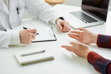 Hastanedeki randevu sırasında bir kadın hastayla bir doktorun ellerinin yakın bir fotoğrafı. İçinde kalem, dosya, dizüstü bilgisayar olan bir defter doktorun ofisinde beyaz bir masada yatıyor..