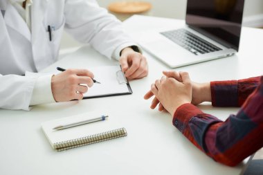Hastanedeki bir randevu sırasında doktorun yanında el kol hareketi yapan bir hastanın katlanmış ellerinin fotoğrafı. İçinde kalem, dosya, dizüstü bilgisayar olan bir defter doktorun ofisinde beyaz bir masada yatıyor.