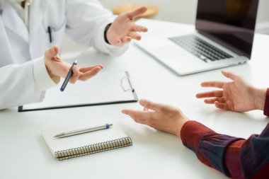 Hastanedeki bir randevu sırasında bir doktorun yanında el kol hareketi yapan bir kadın hastanın yakın bir fotoğrafı. İçinde kalem, dosya, dizüstü bilgisayar olan bir defter doktorun ofisindeki bir masada yatıyor.